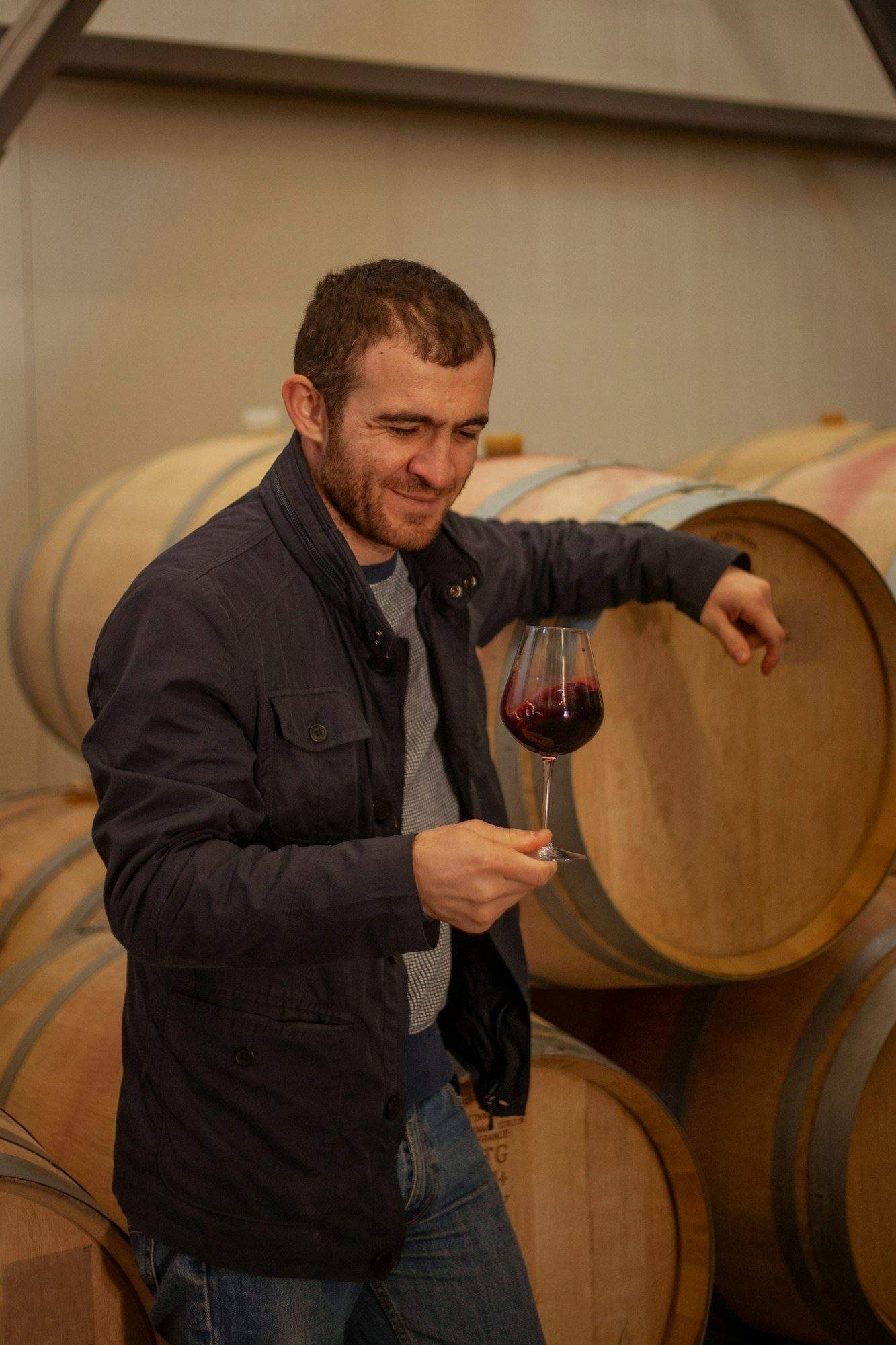 a man winemaker stands near wine barrels and tastes red wine from a glass